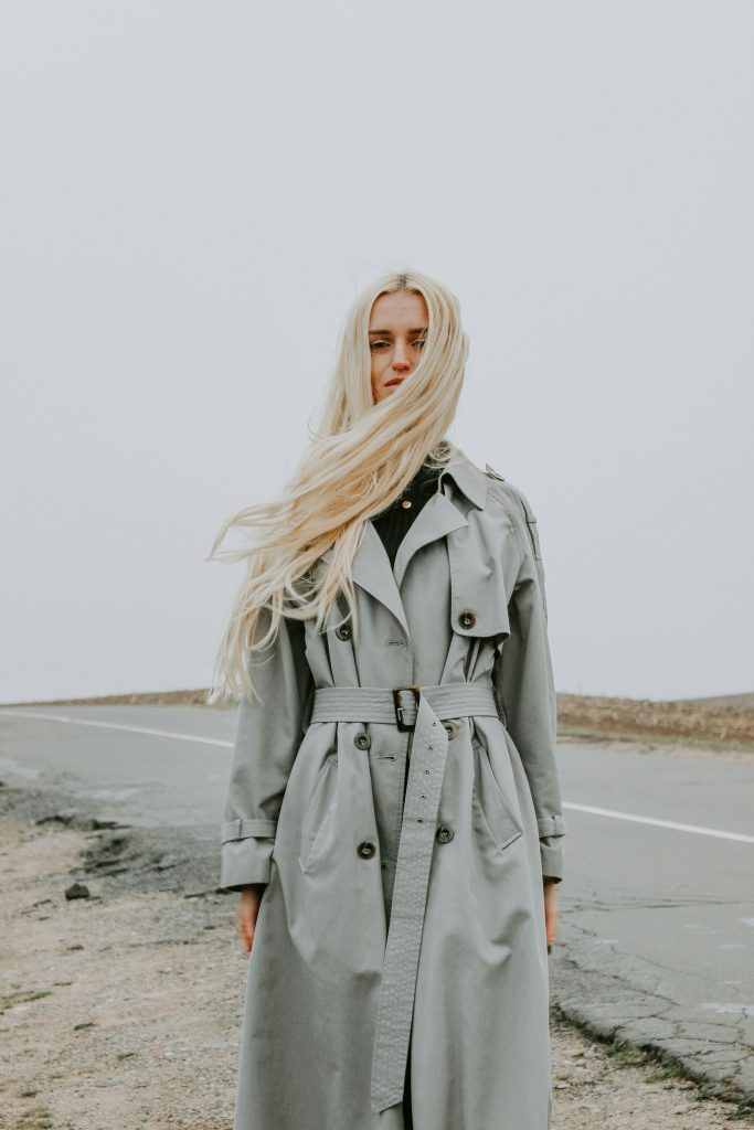 A fashionable woman with long hair poses in a gray coat on a rural road, under an overcast sky.