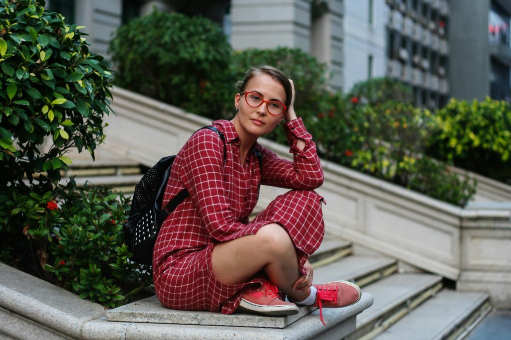 A young woman wearing a red plaid dress and glasses sits on outdoor stairs surrounded by greenery.