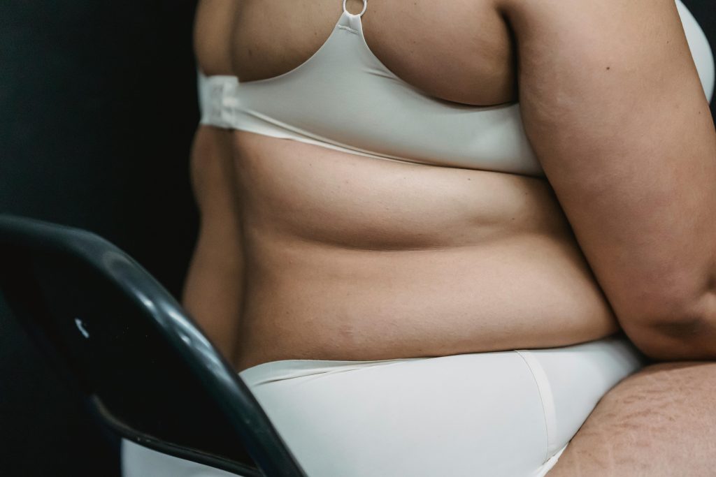 Crop faceless plump woman in white underwear sitting on chair in light studio on dark background