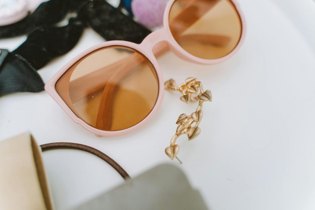 Close-up of trendy pink sunglasses with gold leaf earrings on a white surface. Fashion accessory still life.