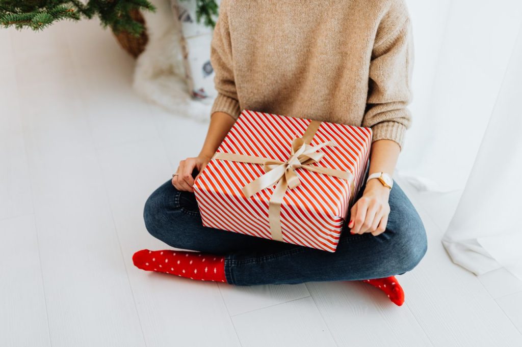 A woman in festive attire holds a beautifully wrapped Christmas gift indoors.