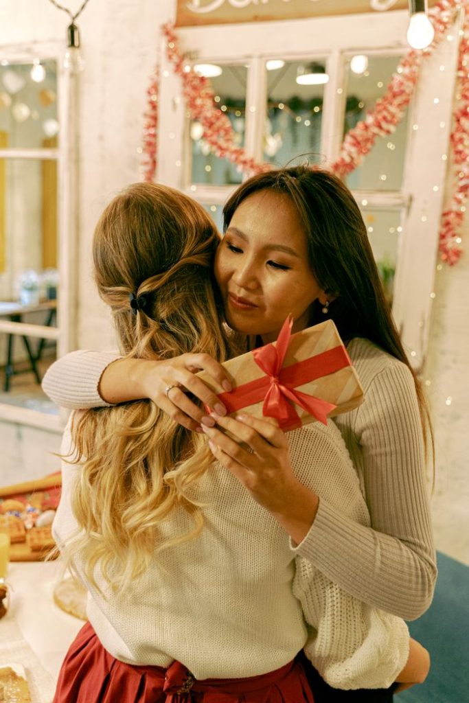 Two women embracing warmly indoors exchanging gifts during Christmas with festive background.